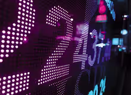 A close-up of a digital display with brightly lit numbers in various shades of purple and blue, possibly representing stock market data or financial information. The background is slightly out of focus, with blurry lights and neon-colored signs in an urban setting.