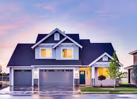 A modern two-story house with white and blue exterior, three-car garage, and a teal front door. Warm interior lights are on, and a small tree is planted in the front yard. The sky is colorful, indicating either sunrise or sunset. The driveway is slightly wet.