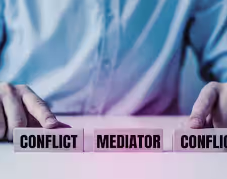 A person in a suit is seen from the torso down, pointing at three wooden blocks on a table. The blocks are labeled "CONFLICT," "MEDIATOR," and "CONFLICT." The image symbolizes the role of a mediator in resolving conflicts between two parties.