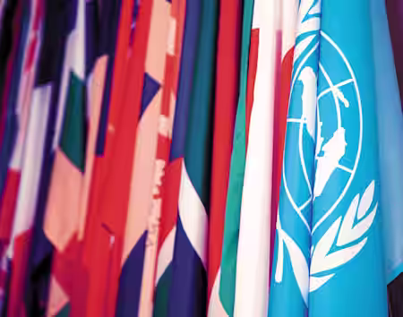 A collection of various national flags is displayed, prominently featuring the blue flag of the United Nations with its white emblem. The flags are closely arranged, showcasing a colorful and diverse array.