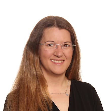 A person with long, straight brown hair, wearing glasses and a black top, is smiling at the camera against a white background.