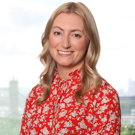 A woman with wavy, shoulder-length blonde hair smiles at the camera. She is wearing a red blouse with a white floral pattern and black pants, standing in front of a window with a blurred cityscape and greenery visible in the background.