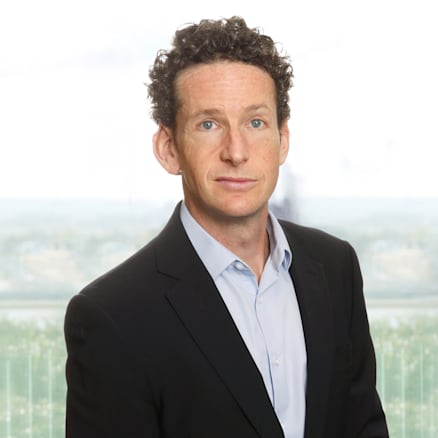 A man with curly hair wearing a light blue collared shirt and a dark blazer stands in front of a blurred background of a window view. He faces forward with a neutral expression.