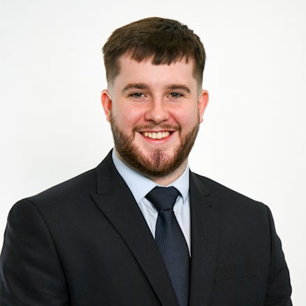 A man with short brown hair and a beard is smiling while wearing a dark suit, light blue dress shirt, and dark tie. He is standing against a plain white background.