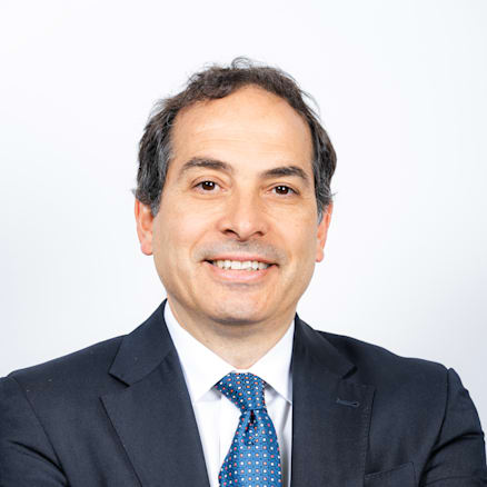 A man in a dark suit with a white dress shirt and a blue patterned tie stands with his arms crossed. He has short, dark hair and is smiling. The background is plain white. He is also wearing a wristwatch on his left hand.