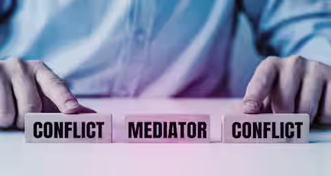 A person in a suit is seen from the torso down, pointing at three wooden blocks on a table. The blocks are labeled "CONFLICT," "MEDIATOR," and "CONFLICT." The image symbolizes the role of a mediator in resolving conflicts between two parties.