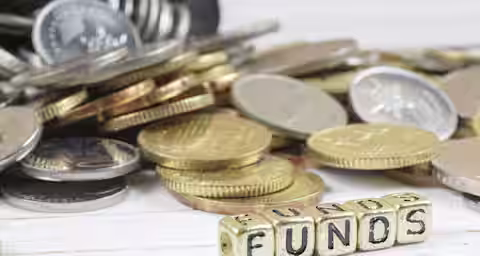 Close-up of various coins stacked and scattered on a wooden surface with gold letter blocks spelling "FUNDS" in the foreground.