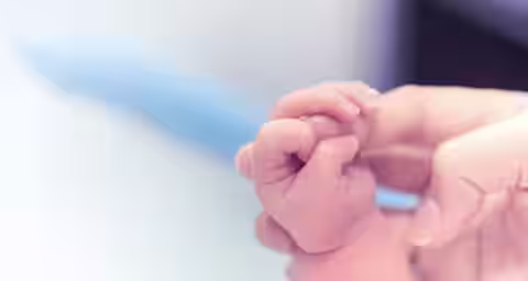 Close-up of an adult's hand gently holding a newborn's tiny hand. The image highlights the tenderness and connection between the adult and the baby. The background is softly blurred, focusing attention on the hands.