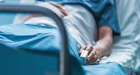 A hospital patient in a blue gown lies in a bed, partially covered by a white blanket. The patient's hand is visible, resting on the blanket, with a pulse oximeter clipped to one of their fingers.
