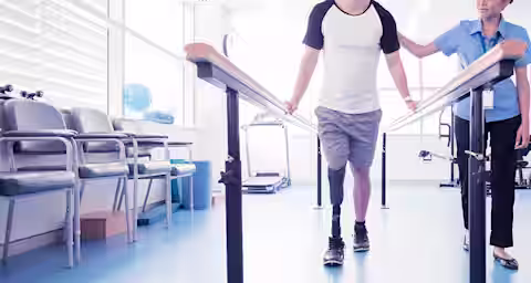 A man with a prosthetic leg is walking between parallel bars in a rehabilitation center while a female therapist supports him. The room is bright with modern equipment and chairs lined up against the wall.