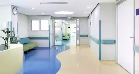 A modern, brightly lit hospital hallway with blue and beige flooring, light green walls, and white doors. There are green cushioned chairs and plants along the left wall. The corridor leads to another section of the hospital through glass doors.