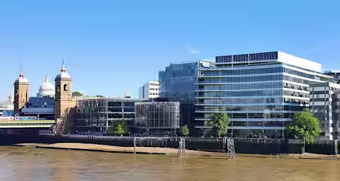 A view of modern and historical buildings lining the River Thames in London, featuring contemporary glass structures alongside traditional architecture with domes. The sky is clear and blue, reflecting on the water below.