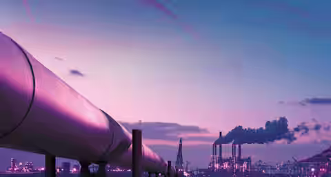 A large oil pipeline extends across a body of water towards an industrial complex with smokestacks emitting smoke. The sky is at dusk, painted in shades of purple and pink, with the water reflecting the colors and lights of the structures.