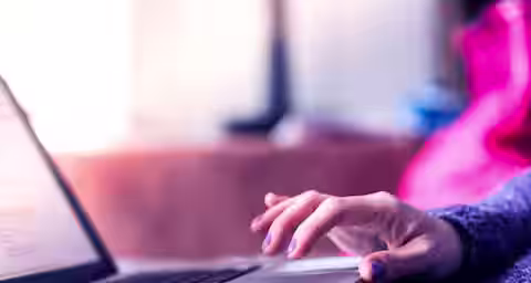 Close-up of a person's hand with purple nail polish resting on the trackpad of an open laptop. The background is softly blurred with pink and blue tones. The person appears to be using the laptop, which is situated on a flat surface.