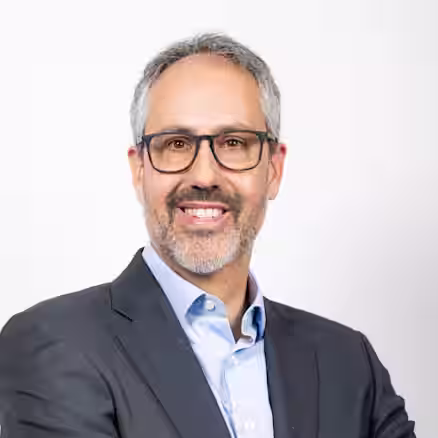 A man with short grey hair and a beard is smiling. He is wearing black-framed glasses, a light blue collared shirt, and a dark suit jacket. His arms are crossed in front of his chest, and he is standing against a plain white background.