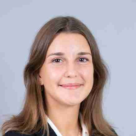 A woman with long brown hair, wearing a black suit jacket over a white blouse, stands against a plain gray background. She is smiling slightly and facing the camera with her hands resting at her sides.