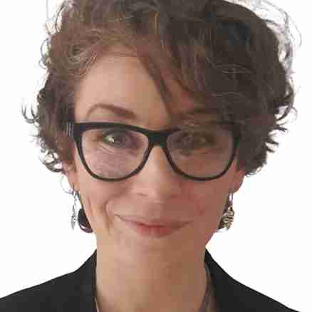 A person with short curly hair and glasses smiles at the camera. They are wearing a black outfit with a large circular pendant necklace and earrings. The background is plain white.