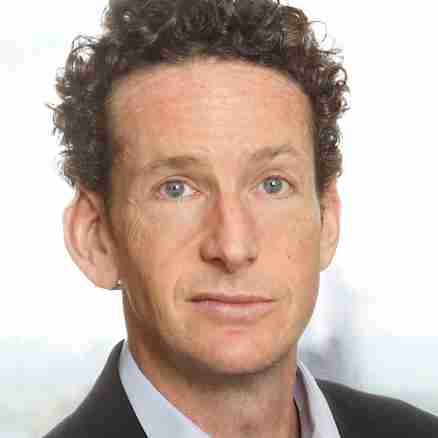 A man with curly hair wearing a light blue collared shirt and a dark blazer stands in front of a blurred background of a window view. He faces forward with a neutral expression.
