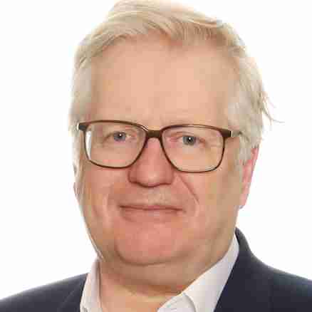 A middle-aged man with light hair and glasses is wearing a dark blazer over a white shirt. He is smiling slightly and standing in front of a plain white background.