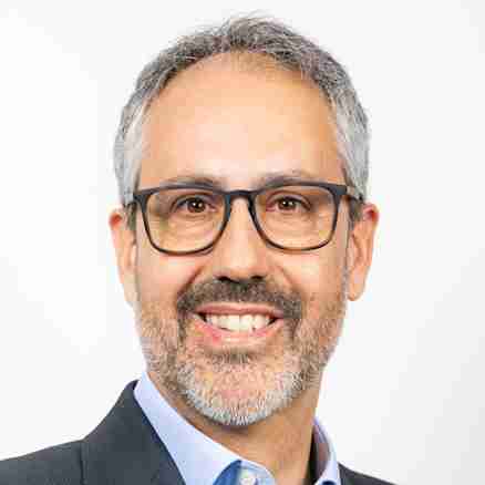A man with short grey hair and a beard is smiling. He is wearing black-framed glasses, a light blue collared shirt, and a dark suit jacket. His arms are crossed in front of his chest, and he is standing against a plain white background.