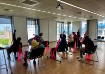 group of people doing yoga sat on chairs, using pink resistance bands