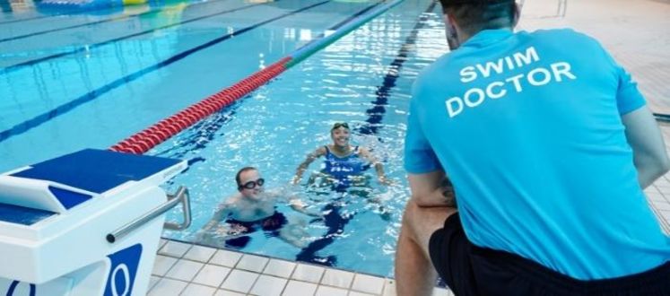 A swimming instructor crouching on the side of the pool, speaking to two people in one of
