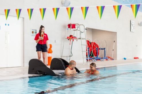 This picture shows a male entering the swimming pool using the pool pod, supported by a female who is in the water and a lifeguard who stands on poolside. 