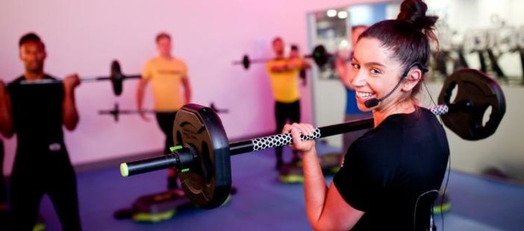 The image shows an instructor smiling at the camera whilst she holds a barbell weight taking a strength fitness class The image shows an instructor smiling at the camera whilst she holds a barbell weight taking a strength fitness class