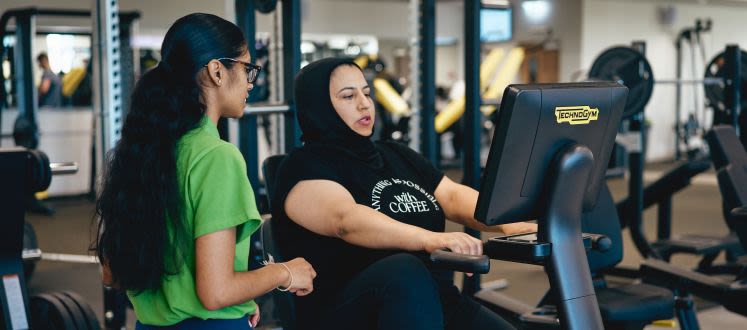 image shows a woman on cardio equipment in a gym with an instructor supporting