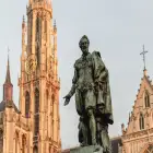A Statue of a Man Wearing a Cape and Extending his Hand Standing in Front of a Gothic Style Clock Tower and Church