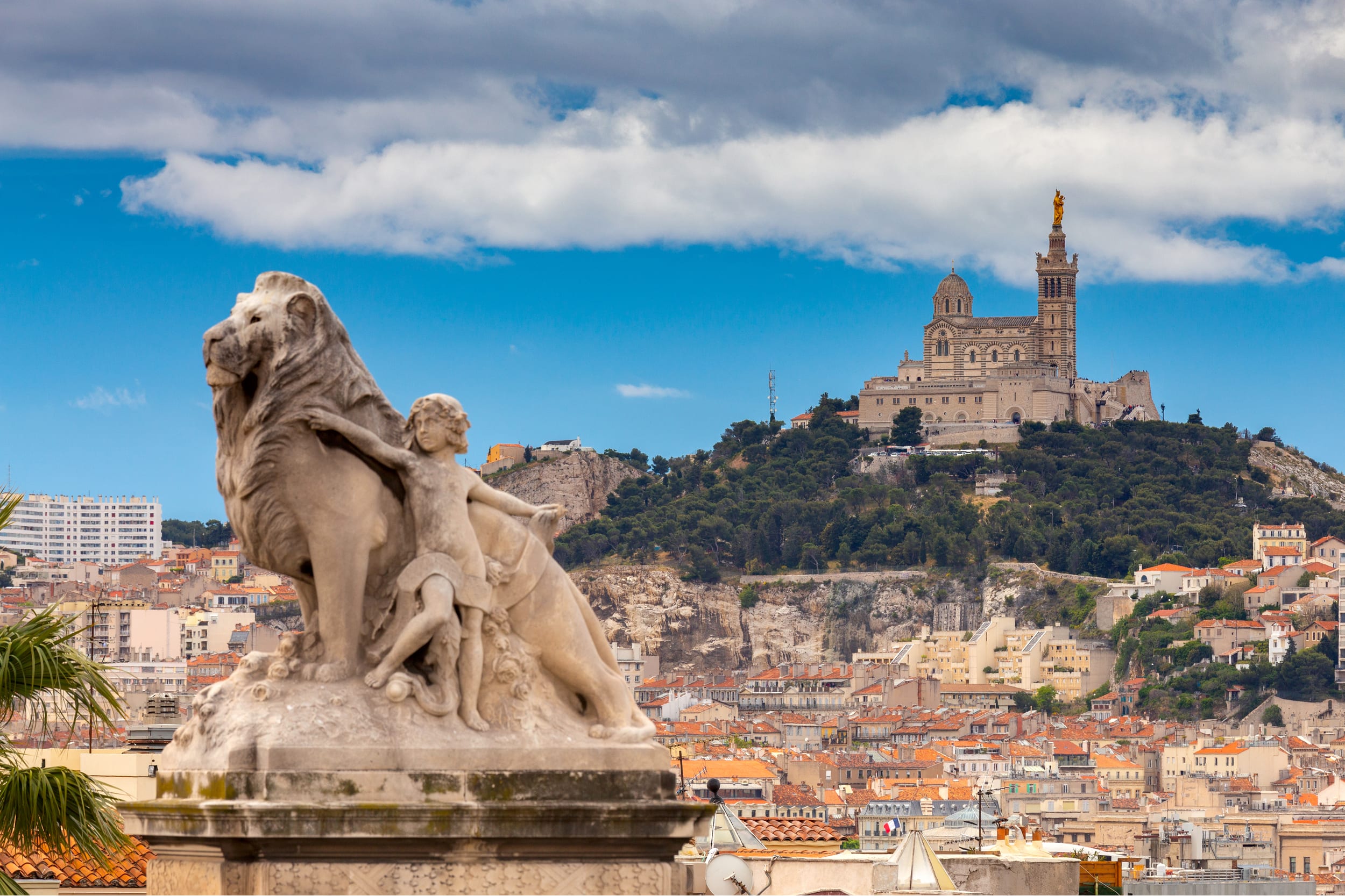 Lion statue from Gare de Marseille-Saint-Charles in the foreground with Basilique Notre-Dame de la Garde and the city of Marseille in the back