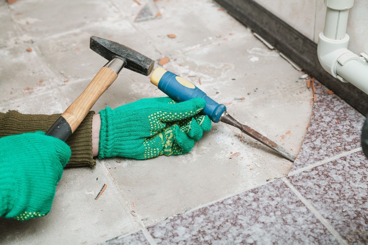 Handwerker entfernt Bodenfliese mit Hammer und Meißel, geschützt durch grüne Handschuhe.
