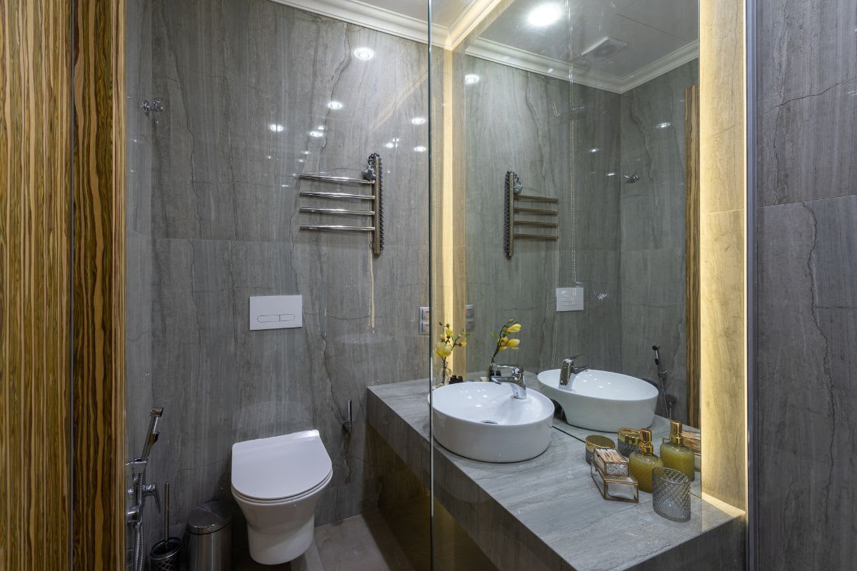 Ein modern gestaltetes Gäste-WC mit grauen Fliesen, einer gläsernen Dusche, einem Doppelwaschbecken und einer spiegelnden Oberfläche, die das Licht reflektiert und für eine offene Raumatmosphäre sorgt.