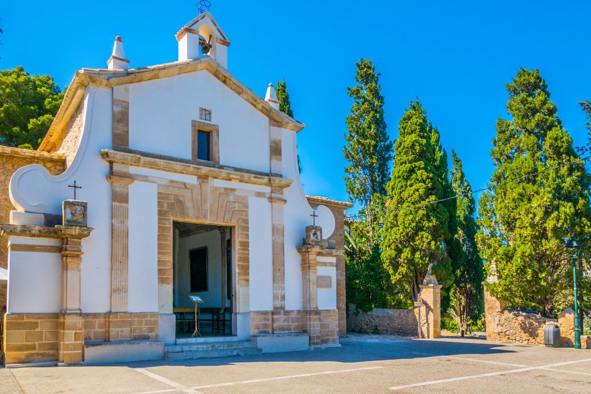 Malerische Kirche auf Mallorca, umgeben von grünen Bäumen und blauem Himmel.