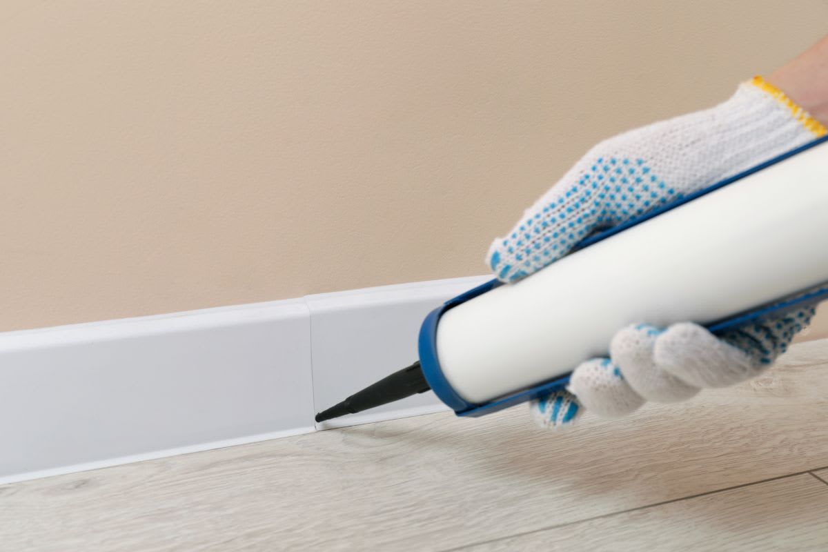 Hand trägt Acryldichtstoff auf die Naht zwischen Sockelleiste und Wand auf.
