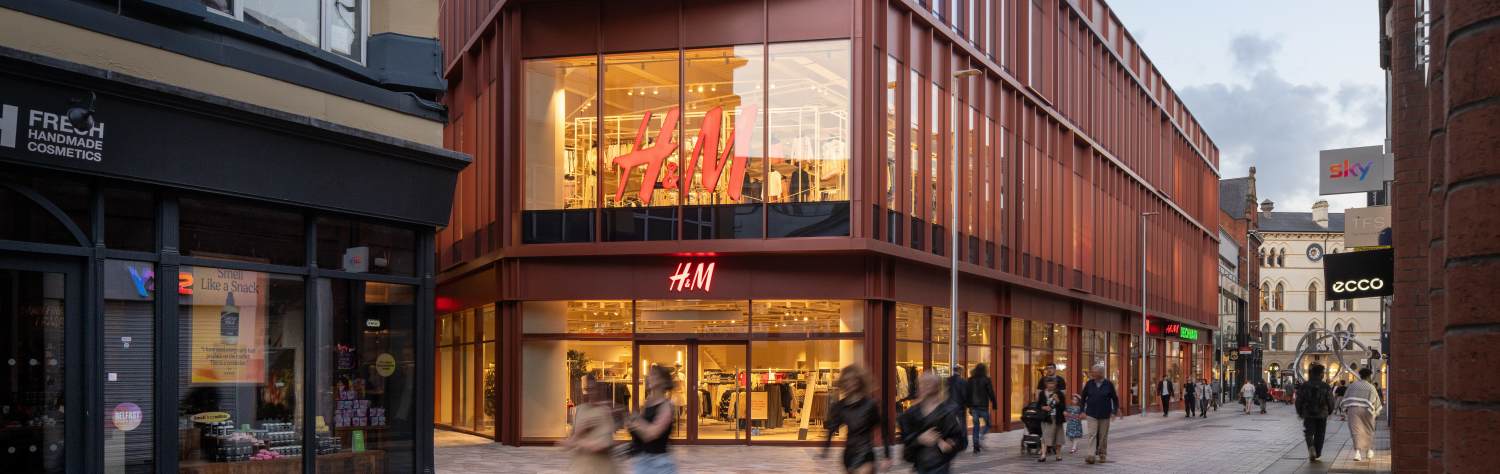 A modern H&M store with a reddish-brown facade and large windows, situated on a bustling city street.