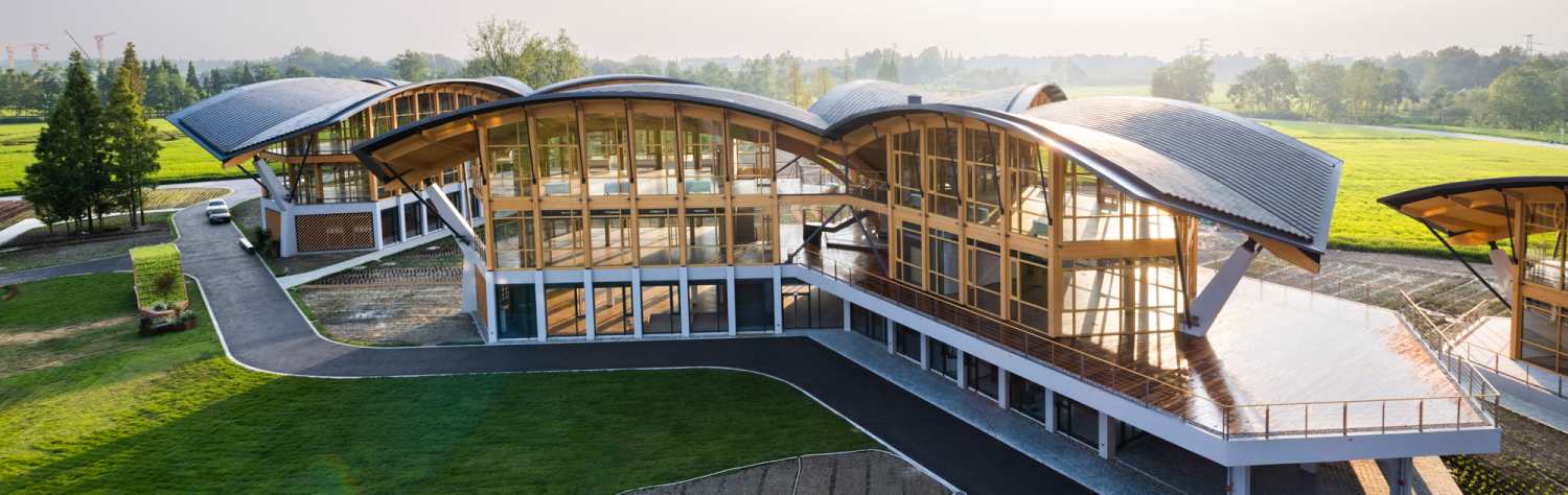 Modern building with curved wooden roofs and large glass walls set in a green landscape.