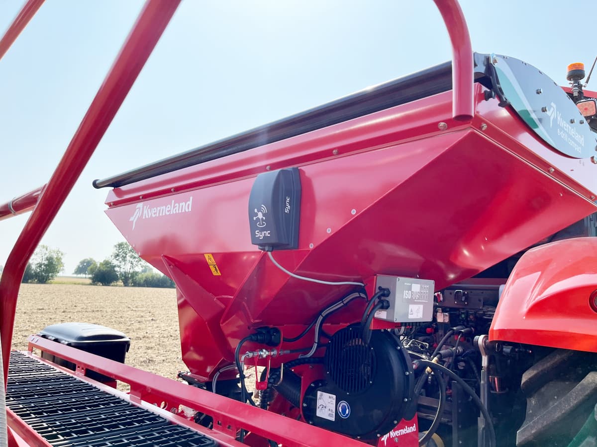 Close-up of the small black Kverneland Sync module mounted on a red Kverneland fertilizer spreader spreader, showing its placement and wiring in a sunlit field