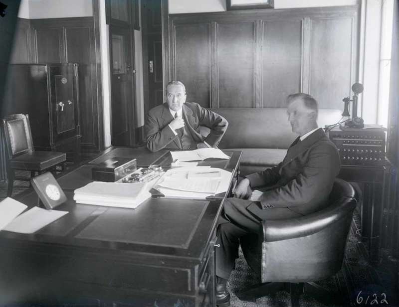 This is a black-and-white photograph showing Prime Minister Stanley Bruce and James Scullin seated at a desk in the prime minister's office in (Old) Parliament House, the day before Scullin was sworn in as the new Australian prime minister. Scullin is seated in a leather chair in front of the desk and Bruce is seated to his right at the side of the desk. Papers and writing materials cover much of the desk and a telephone is visible in the background. The room is wood panelled.