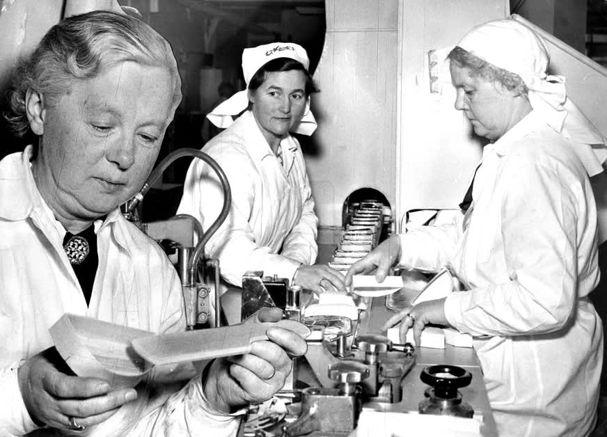 Sort og hvitt-foto av osteemballasje på løpende bånd i den tidligere fabrikken til Kavli på Damsgård. Ved båndet inspiseres osteeskene nøye av Kavli-medarbeidere i hvit uniform.