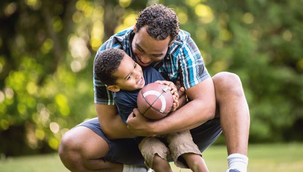 Cute African American father and son smiling in an outdoor park - lots of green background. Looking at camera and happy.Cute African American father and son smiling in an outdoor park - lots of green background. Playing Football