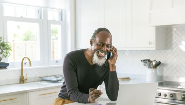 A smiling middle aged Black man speaks on the telephone in a sunny kitchen.