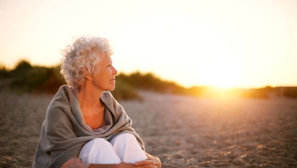 person looking concerned or deep in thought while sitting on the beach with the sun setting behind them