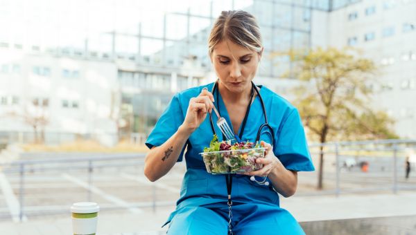 a white woman nurse in blue scrubs sits outside during her lunch break eating a fresh salad 