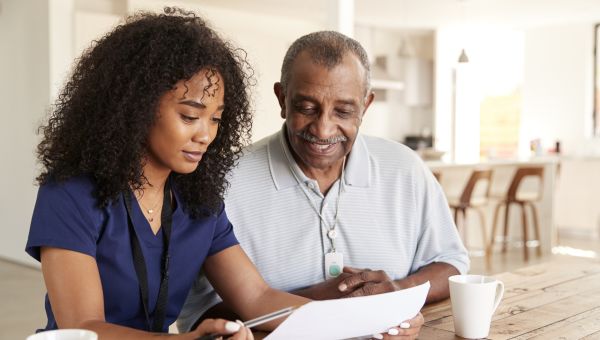 a young Black nurse sits at a dining room table with an older Black male patient going over his medications and treatment plan