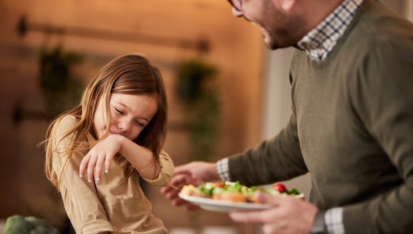 girl with autism sits on the dining table laughing with her dad holding a plate of food trying to cheerfully encourage her to eat