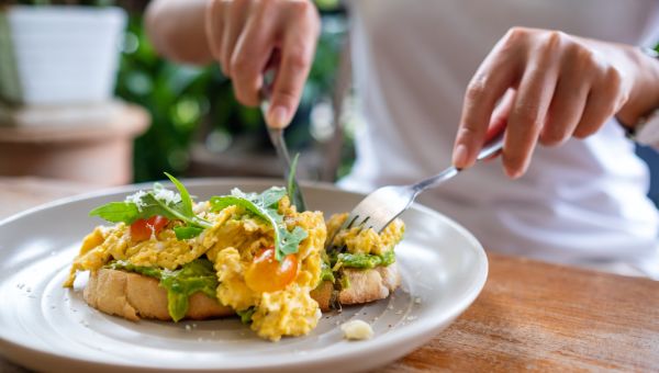 closeup of a plate of scrambled eggs over toast with arugula while an unidentified woman's hands uses a fork and knife to cut into it