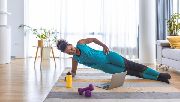 young woman on a rug on her floor doing a side plank while watching an fitness video on her laptop