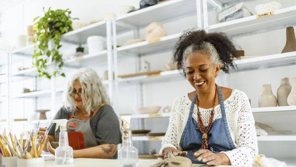 a Black and white woman sit side by side at a table at an art class, creating ceramic art