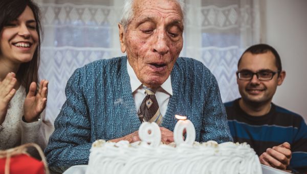 an older white man celebrates his 90th birthday with loved ones; he sits in front of a birthday cake and blows out the candles